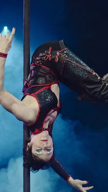 ADM Spectacles - Une personne, en pose acrobatique, est suspendue la tête en bas à un poteau, tel un spectacle éblouissant. Elle porte une tenue noire et rouge ornée de paillettes. Une fumée bleue emplit l'arrière-plan, et l'expression de son visage est saisissante lors de ces spectacles.