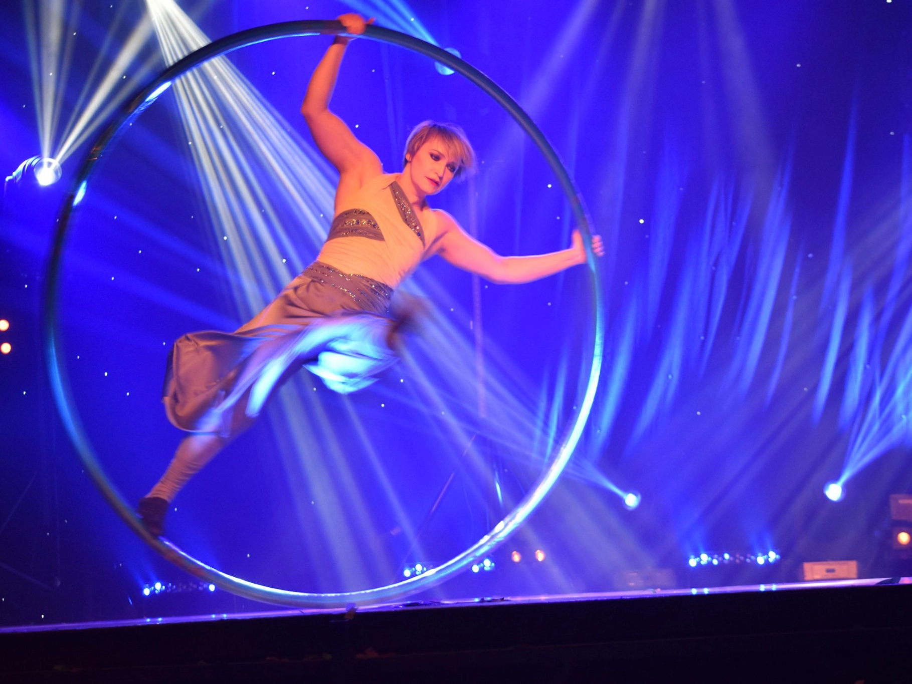 ADM Spectacles - Un artiste aux cheveux blonds courts évolue avec habileté à l'intérieur d'un grand cerceau circulaire sur scène. La scène, qui fait partie intégrante de ce spectacle éblouissant, est illuminée par des lumières bleues et violettes vibrantes, créant une atmosphère dynamique et énergique.