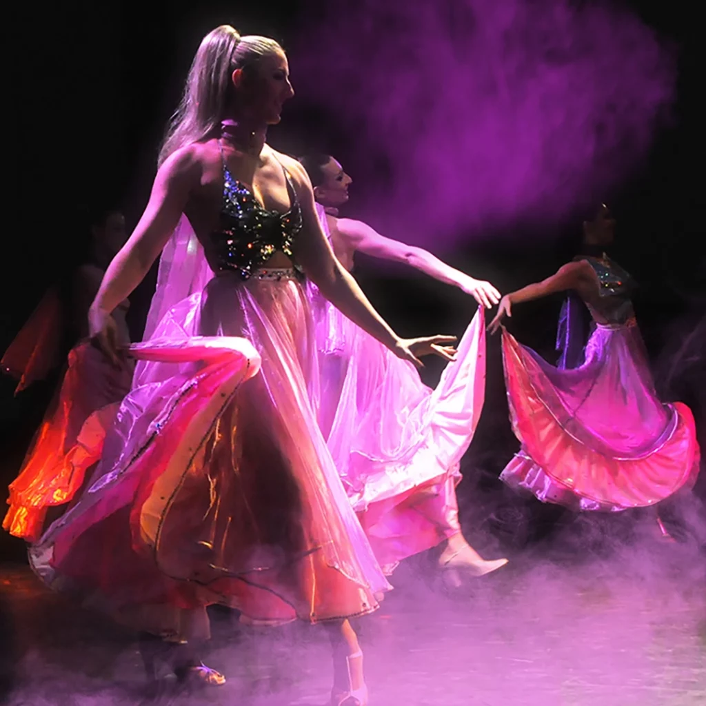 ADM Spectacles - Des danseuses vêtues de robes lavande fluides et brillantes se produisent sur scène, entourées de fumée rose et violette. Leurs costumes scintillent sous les projecteurs, tels de véritables artistes, créant un spectacle de mouvements gracieux et synchronisés.