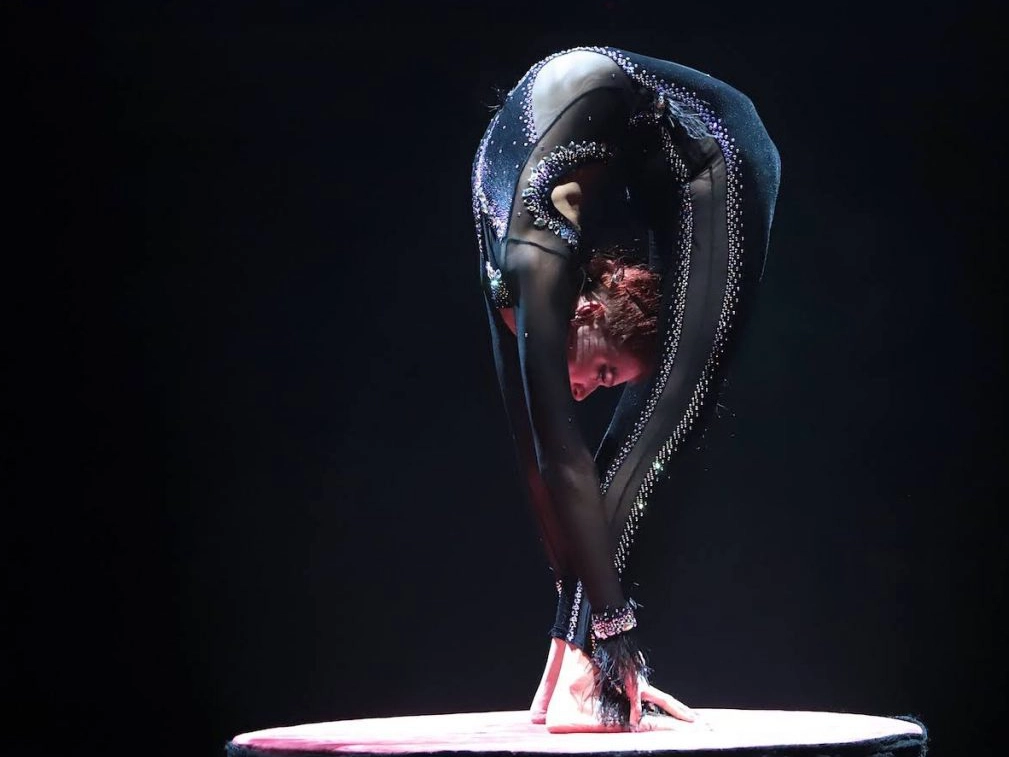 ADM Spectacles - Un contorsionniste vêtu d'une combinaison noire moulante aux détails scintillants exécute une flexion arrière extrême sur une plateforme circulaire. Tels des danseurs expérimentés, leur corps est penché en arrière, mains et pieds touchant la surface. La lumière met en valeur leur souplesse et leur équilibre.