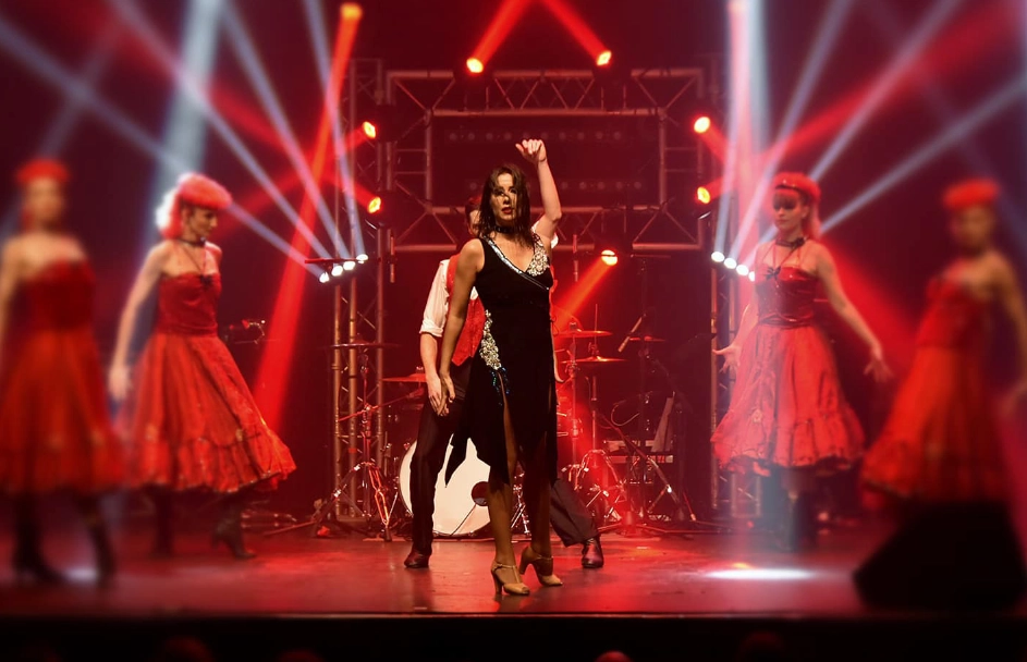 ADM Spectacles - Un groupe d'artistes monte sur scène, avec une femme en tenue noire dansant au centre. Autour d'elle, des danseurs en robes rouges évoluent avec grâce. L'atmosphère théâtrale vibrante est sublimée par des projecteurs rouges et blancs, créant un événement ADM Spectacles inoubliable.